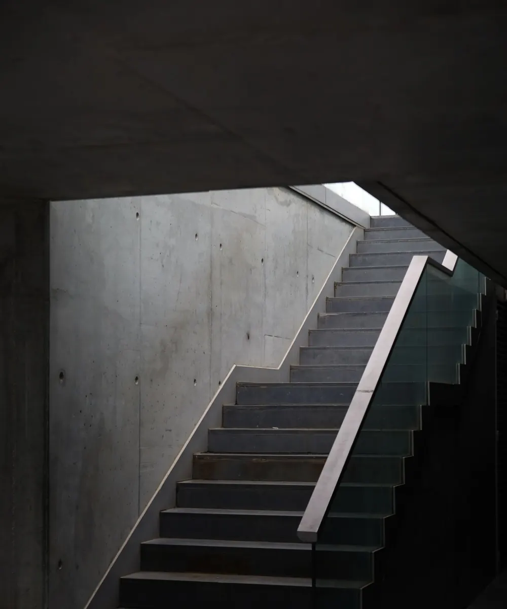 Cage d'escalier en béton banché montant vers la lumière du jour, main courante d'acier.