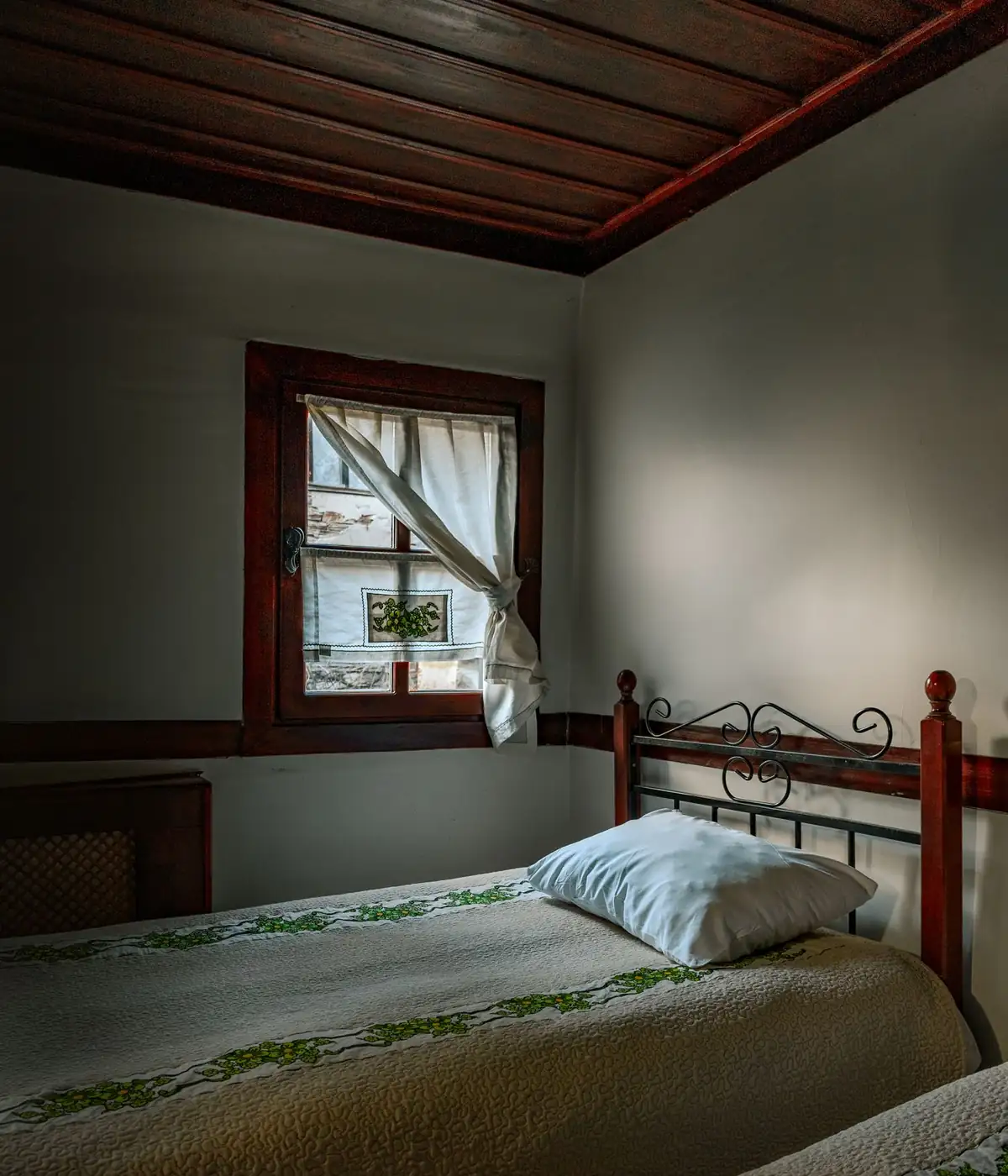 Chambre aux murs clairs et plafond de planches sombres : lit de fer, couvre-lit blanc brodé de feuillage vert, fenêtre à petits carreaux et rideau de lin retenu.