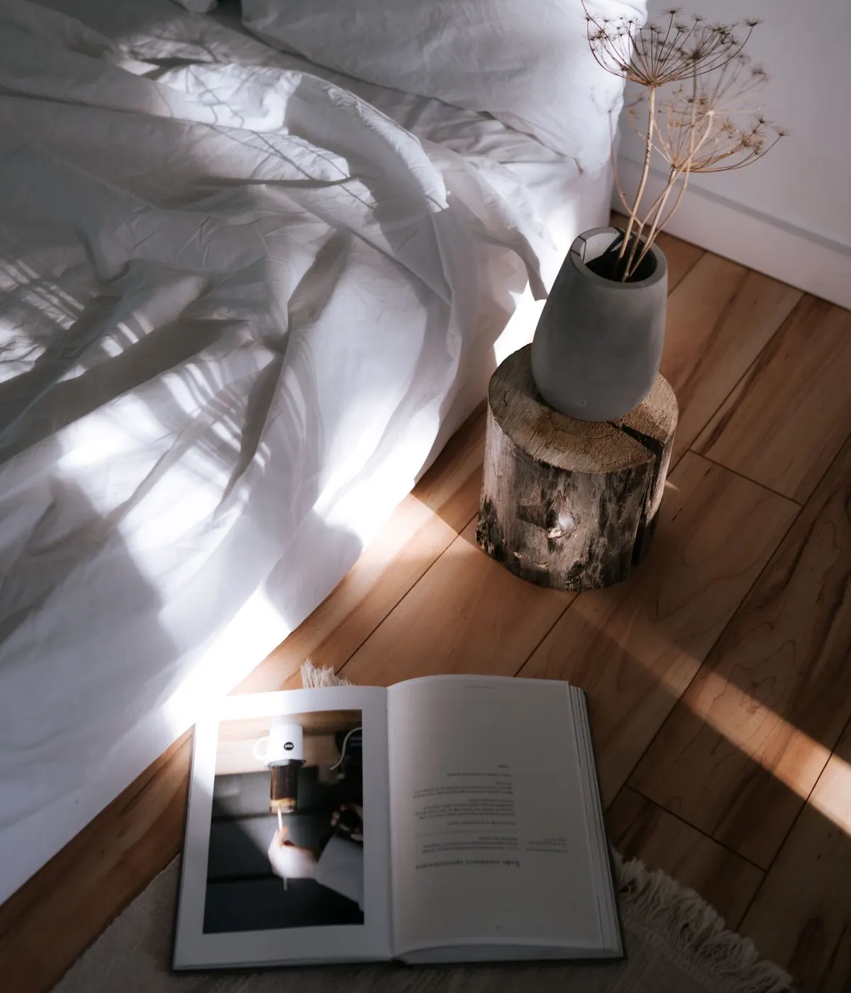 Coin de chambre au petit matin : draps de lin blanc froissés, tabouret en rondin portant un vase de fleurs séchées, un livre ouvert posé sur un tapis à franges.
