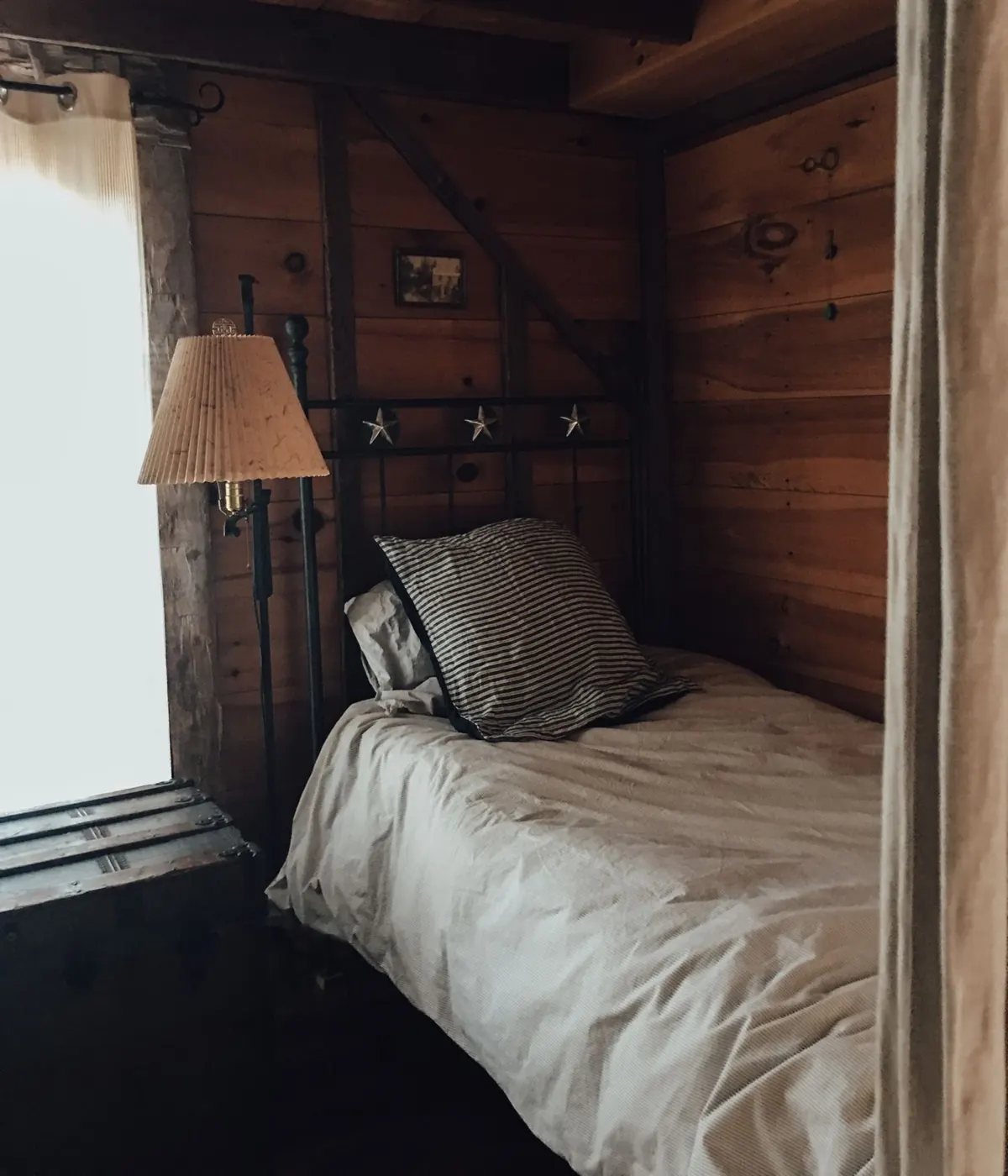 Petite chambre lambrissée de bois brun : lit une place à cadre de fer, édredon écru, coussin à rayures, lampadaire à abat-jour plissé et vieille malle sous la fenêtre.