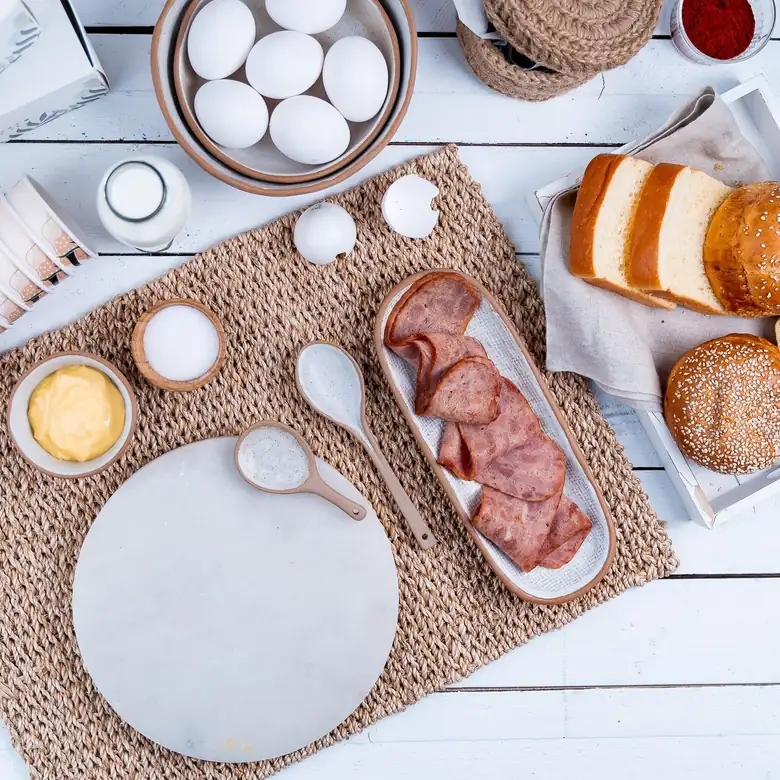 Petit-déjeuner en préparation, vu de dessus : œufs dans un bol, bouteille de lait, brioche et petits pains tranchés, planche de marbre et ramequins sur un set tressé.