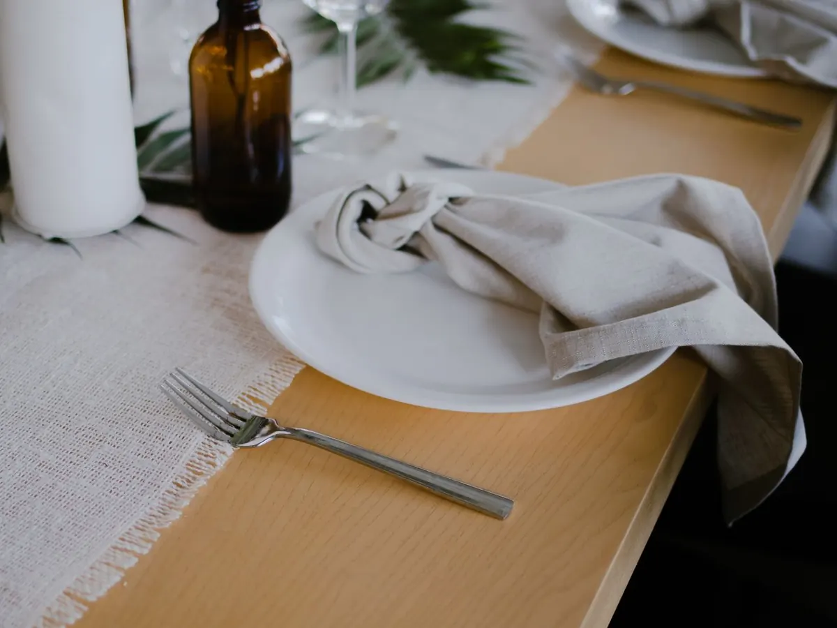 Couvert dressé : assiette blanche, serviette de lin nouée, fourchette posée sur le bois nu, chemin de table tissé et bouteille en verre ambré.